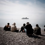 Greece, Patras. A group of refugees sit on a beach as they watch an approaching ship.