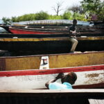 On a beach near the port, a young man uses a ruler to carefully draw out the name of a refurbished fisherman's canoe onto its side.