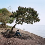 A man wait sitting in the shadow of a tree.